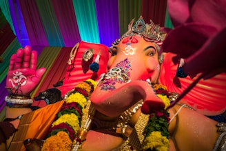 A vibrant statue of Lord Ganesha is adorned with intricate jewelry and colorful garlands. The statue's trunk is decorated with artistic patterns and it holds a lotus in its hand. The backdrop features a multicolored draped fabric, with an overall festive and energetic atmosphere.