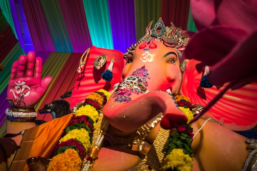 A vibrant statue of Lord Ganesha is adorned with intricate jewelry and colorful garlands. The statue's trunk is decorated with artistic patterns and it holds a lotus in its hand. The backdrop features a multicolored draped fabric, with an overall festive and energetic atmosphere.