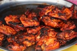 A plate filled with pieces of grilled chicken, cooked to a crispy texture with a rich, reddish-brown color. The chicken appears to be marinated with spices, leading to a charred and smoky appearance.