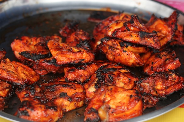 A plate filled with pieces of grilled chicken, cooked to a crispy texture with a rich, reddish-brown color. The chicken appears to be marinated with spices, leading to a charred and smoky appearance.