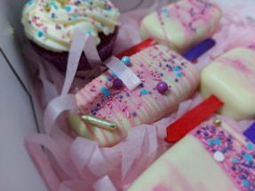 A box containing elegantly decorated desserts, including cake popsicles coated in white chocolate with pink drizzle and colorful sprinkles, alongside a frosted cupcake adorned with similar sprinkles. The treats are nestled in pink shredded paper, creating a festive and inviting presentation.