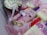 A box containing elegantly decorated desserts, including cake popsicles coated in white chocolate with pink drizzle and colorful sprinkles, alongside a frosted cupcake adorned with similar sprinkles. The treats are nestled in pink shredded paper, creating a festive and inviting presentation.
