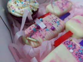 A box containing elegantly decorated desserts, including cake popsicles coated in white chocolate with pink drizzle and colorful sprinkles, alongside a frosted cupcake adorned with similar sprinkles. The treats are nestled in pink shredded paper, creating a festive and inviting presentation.