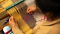 A person is weaving colorful patterns on a traditional loom. The textile being woven features geometric shapes in vibrant shades of blue, purple, and red. Their hands skillfully manipulate the threads, creating intricate designs. Wooden rods and warp threads are visible, showcasing the detailed weaving process.