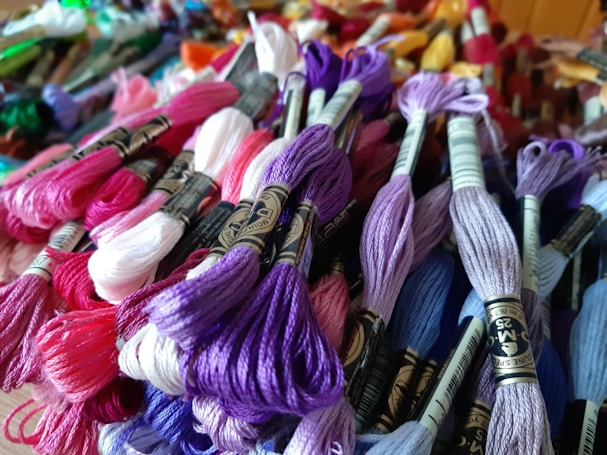 A variety of colorful embroidery threads are tightly packed together, showcasing an array of hues such as purple, pink, lavender, and white. The threads are skeins, each wrapped with a paper band bearing labels. The background contains a blur of more threads and other crafting items, adding to the vibrant and busy scene.