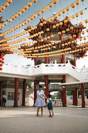 A traditional Chinese pavilion is adorned with numerous yellow lanterns that hang in decorative rows against a clear sky. In the foreground, an adult in a blue dress interacts joyfully with a child in overalls, surrounded by a spacious plaza with a white railing and red pillars.