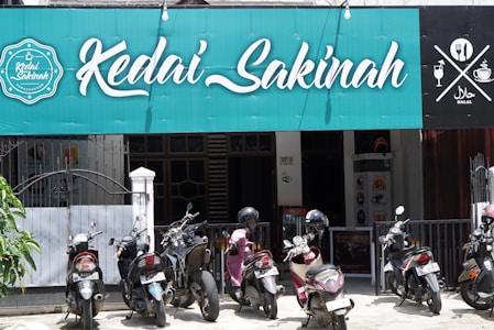 A storefront with a teal sign that reads 'Kedai Sakinah.' Several motorcycles are parked in front of the establishment. To the right of the sign, there are symbols indicating that the place is halal and has food and drink offerings. The frontage includes a gate and pillars.