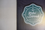 A textured wall features a logo for 'Kedai Sakinah Panakkukang.' The logo is circular with a teal background and white text and design elements. It includes a small coffee cup icon above stylized script writing, surrounded by coffee bean symbols.