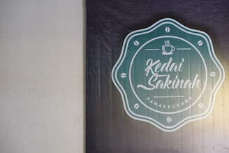 A textured wall features a logo for 'Kedai Sakinah Panakkukang.' The logo is circular with a teal background and white text and design elements. It includes a small coffee cup icon above stylized script writing, surrounded by coffee bean symbols.