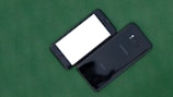 Two smartphones are placed side by side on a green surface. One phone is face down showing the brand 'Samsung,' while the other is face up with a blank white screen. Both phones are black, with sleek and shiny finishes.