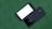 Two smartphones are placed side by side on a green surface. One phone is face down showing the brand 'Samsung,' while the other is face up with a blank white screen. Both phones are black, with sleek and shiny finishes.