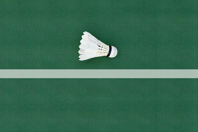 A single badminton shuttlecock rests on a green court with a white line running horizontally in the background. The shuttlecock is centered and its white feathers and cork tip are clearly visible.