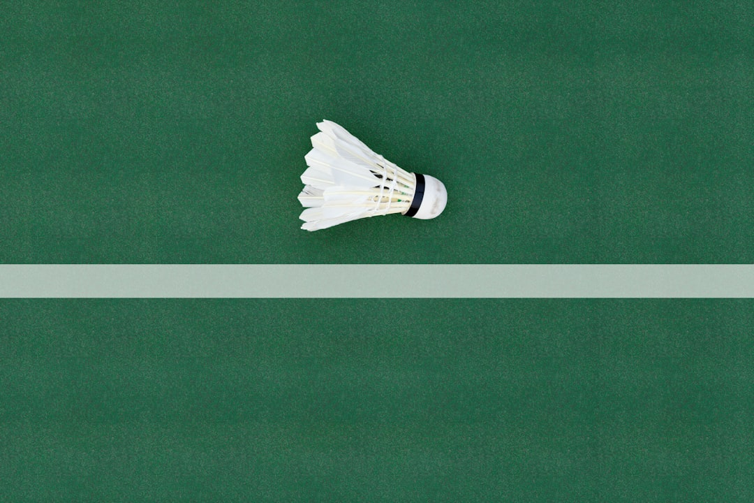white and black shoe on green surface, A white badminton feather shuttlecock at a corner on a badminton court.