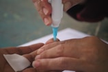 A close-up of hands using a small medical device with a blue tip to perform a procedure on a fingertip. A cotton pad is held nearby, suggesting a medical or testing context.