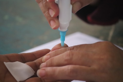A close-up of hands using a small medical device with a blue tip to perform a procedure on a fingertip. A cotton pad is held nearby, suggesting a medical or testing context.