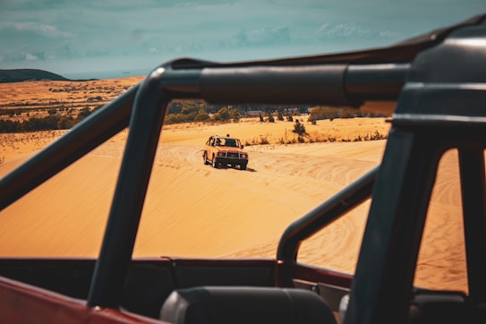 A jeep is navigating through a sandy desert landscape with dunes extending into the distance. The foreground features the frame of another vehicle, indicating a possible off-road adventure. Sparse vegetation can be seen in the background, adding to the arid setting.
