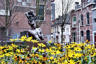 A bronze statue of a woman with an abstract face mask in a city courtyard is surrounded by vibrant yellow flowers. In the background, brick residential buildings with windows and some leafless trees can be seen. The scene conveys a blend of nature and urban art.