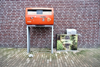 A red post box stands against a brick wall, decorated with stickers and logos. To its right, there is a metal box covered with an image of a serene nature scene featuring a stream and greenery. The ground is paved with bricks, and a crumpled paper lies in front of the post box.
