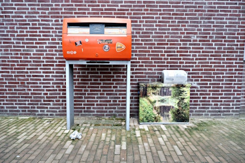 A red post box stands against a brick wall, decorated with stickers and logos. To its right, there is a metal box covered with an image of a serene nature scene featuring a stream and greenery. The ground is paved with bricks, and a crumpled paper lies in front of the post box.