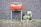 A red post box stands against a brick wall, decorated with stickers and logos. To its right, there is a metal box covered with an image of a serene nature scene featuring a stream and greenery. The ground is paved with bricks, and a crumpled paper lies in front of the post box.