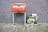A red post box stands against a brick wall, decorated with stickers and logos. To its right, there is a metal box covered with an image of a serene nature scene featuring a stream and greenery. The ground is paved with bricks, and a crumpled paper lies in front of the post box.