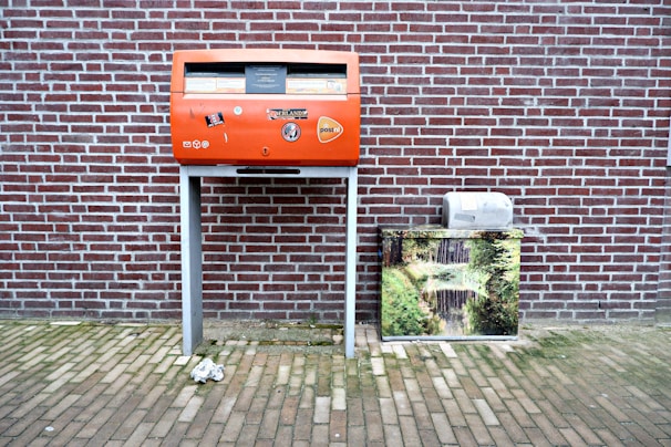A red post box stands against a brick wall, decorated with stickers and logos. To its right, there is a metal box covered with an image of a serene nature scene featuring a stream and greenery. The ground is paved with bricks, and a crumpled paper lies in front of the post box.