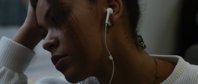 A person wearing white earbuds is leaning their head against their hand, appearing thoughtful or contemplative. The background is blurry, suggesting a close-up focus on the face.