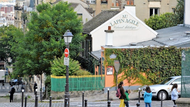 An artistic cabaret building with a sign reading 'Au Lapin Agile' is surrounded by lush greenery and ivy. People are walking along a cobblestone street in front of the building. A classic street lamp and a stop sign add to the quaint urban scene.