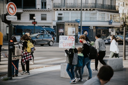 A group of people, including children, are walking across a street in an urban area. One child holds a protest sign with French text related to climate change. The street is busy with cars and there are buildings in the background. Some individuals are wearing backpacks and casual clothing.