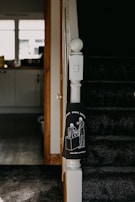 The image captures a section of a staircase in a home, with dark carpeting and a white newel post. Attached to the newel post is a black cloth featuring a white line drawing of two people standing at a table under the phrase 'SUPPORT SMALL BUSINESSES'. The background includes a view of a kitchen area with a counter, cabinets, and items on the counter, lit by natural light from the window.