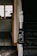 The image captures a section of a staircase in a home, with dark carpeting and a white newel post. Attached to the newel post is a black cloth featuring a white line drawing of two people standing at a table under the phrase 'SUPPORT SMALL BUSINESSES'. The background includes a view of a kitchen area with a counter, cabinets, and items on the counter, lit by natural light from the window.