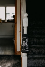 The image captures a section of a staircase in a home, with dark carpeting and a white newel post. Attached to the newel post is a black cloth featuring a white line drawing of two people standing at a table under the phrase 'SUPPORT SMALL BUSINESSES'. The background includes a view of a kitchen area with a counter, cabinets, and items on the counter, lit by natural light from the window.