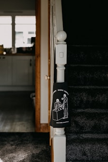The image captures a section of a staircase in a home, with dark carpeting and a white newel post. Attached to the newel post is a black cloth featuring a white line drawing of two people standing at a table under the phrase 'SUPPORT SMALL BUSINESSES'. The background includes a view of a kitchen area with a counter, cabinets, and items on the counter, lit by natural light from the window.