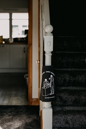 The image captures a section of a staircase in a home, with dark carpeting and a white newel post. Attached to the newel post is a black cloth featuring a white line drawing of two people standing at a table under the phrase 'SUPPORT SMALL BUSINESSES'. The background includes a view of a kitchen area with a counter, cabinets, and items on the counter, lit by natural light from the window.