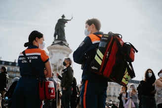 Several people are gathered in an urban outdoor setting. Two individuals in the foreground, wearing uniforms with high-visibility markings, appear to be part of an emergency or medical services team. They wear masks and are engaged in conversation. One person carries a large backpack with emergency supplies. In the background, a statue is situated atop a pedestal, and more people can be seen, also wearing masks, indicating an awareness of health precautions.