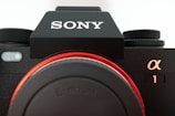 Close-up view of a black Sony camera with a large lens mount and textured control dials. Prominent Sony logo in white on the black body, with the alpha symbol and number one next to it.