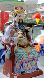 A colorful ceramic figurine of a seated bearded figure wearing ornate traditional attire and a crown, holding a small golden object. The clothing is decorated with intricate patterns in various colors. Surrounding the figurine are ceramic vases with floral designs. Red lanterns and green foliage are visible in the background, suggesting an Asian cultural setting.