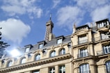 A Parisian building with classic architectural features is in the foreground, with the Eiffel Tower rising prominently in the background against a blue sky with scattered clouds. The sunlight reflects off the building's windows, creating a bright spot.