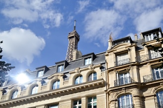 A Parisian building with classic architectural features is in the foreground, with the Eiffel Tower rising prominently in the background against a blue sky with scattered clouds. The sunlight reflects off the building's windows, creating a bright spot.
