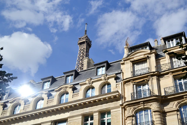 A Parisian building with classic architectural features is in the foreground, with the Eiffel Tower rising prominently in the background against a blue sky with scattered clouds. The sunlight reflects off the building's windows, creating a bright spot.