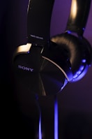 A close-up view of over-ear black headphones with a metallic finish, featuring the brand name and model number in white lettering. The lighting casts soft blue and purple highlights on the headphones, emphasizing their sleek design.