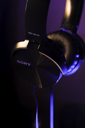 A close-up view of over-ear black headphones with a metallic finish, featuring the brand name and model number in white lettering. The lighting casts soft blue and purple highlights on the headphones, emphasizing their sleek design.