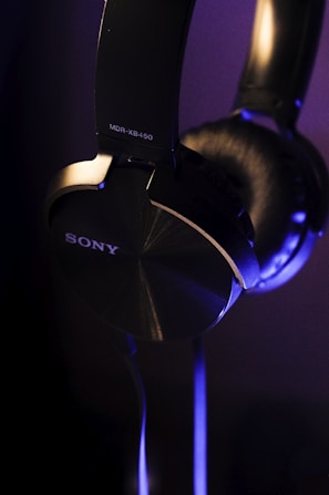 A close-up view of over-ear black headphones with a metallic finish, featuring the brand name and model number in white lettering. The lighting casts soft blue and purple highlights on the headphones, emphasizing their sleek design.