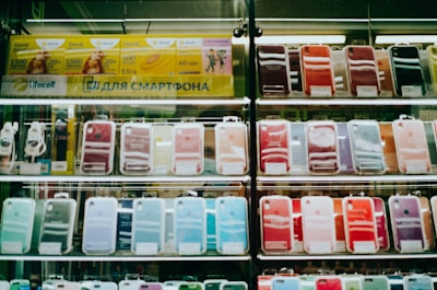 Shelves display a variety of smartphone cases in multiple colors, arranged in an organized manner within a store. A sign in Cyrillic characters and advertisements for mobile network plans are visible above the cases.