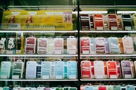 Shelves display a variety of smartphone cases in multiple colors, arranged in an organized manner within a store. A sign in Cyrillic characters and advertisements for mobile network plans are visible above the cases.