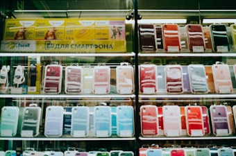 Shelves display a variety of smartphone cases in multiple colors, arranged in an organized manner within a store. A sign in Cyrillic characters and advertisements for mobile network plans are visible above the cases.