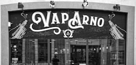 A storefront sign for a shop named 'Vap Arno', featuring stylized text and illustrations of vaping devices on either side. The sign includes the establishment year, 2017, and the storefront has large windows showing some interior elements.