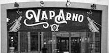 A storefront sign for a shop named 'Vap Arno', featuring stylized text and illustrations of vaping devices on either side. The sign includes the establishment year, 2017, and the storefront has large windows showing some interior elements.
