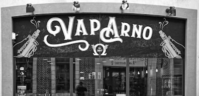 A storefront sign for a shop named 'Vap Arno', featuring stylized text and illustrations of vaping devices on either side. The sign includes the establishment year, 2017, and the storefront has large windows showing some interior elements.