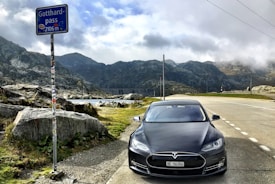 A Tesla car is parked on a mountain pass road with rugged hills in the background. The sky is partly cloudy, casting shadows on the landscape. A metal signpost indicating the Gotthard Pass and its elevation stands nearby.